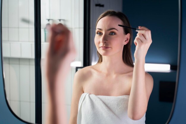 Une jolie jeune femme peint ses cils avec du mascara devant le miroir de la salle de bain