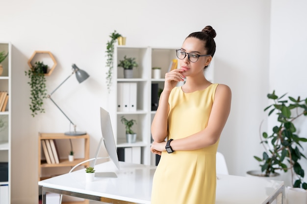 Jolie jeune femme pensive en élégante robe jaune et lunettes debout par lieu de travail au bureau