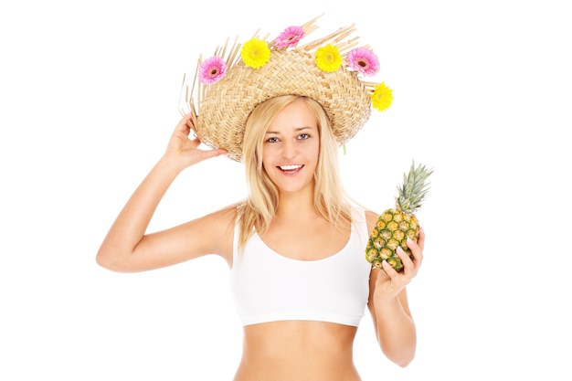 une jolie jeune femme posant dans un chapeau avec des fleurs et un ananas sur fond blanc