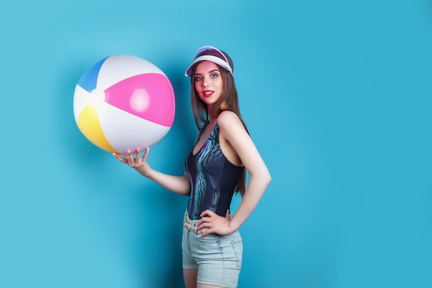 Jolie jeune femme posant en maillot de bain et casquette visière en plastique rose avec ballon gonflable sur fond bleu