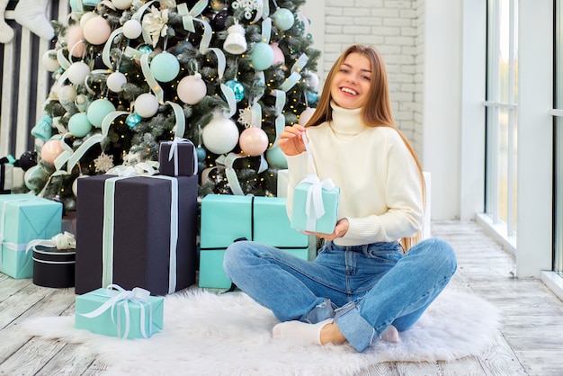 jolie jeune femme près de l'arbre de noël ouvre des cadeaux de noël dans une pièce lumineuse et confortable