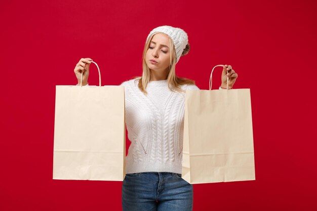 Jolie jeune femme en pull blanc, chapeau isolé sur fond de mur rouge en studio. Mode de vie sain, concept de saison froide. Maquette de l'espace de copie. Tenez le sac d'emballage avec les achats après le shopping.