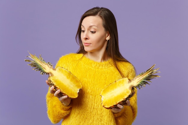 Jolie jeune femme en pull de fourrure tenir dans les mains à la recherche de moitiés de fruits d'ananas mûrs frais isolés sur fond pastel violet. Mode de vie vivant des gens, concept de vacances relaxantes. Maquette de l'espace de copie.