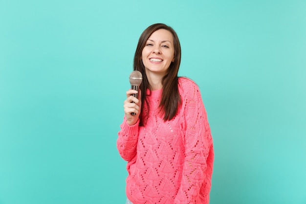 Jolie Jeune Femme En Pull Rose Tricoté Tenant à La Main Et Chanter Une Chanson Dans Un Microphone Isolé Sur Fond De Mur Bleu Turquoise, Portrait En Studio. Concept De Mode De Vie Des Gens. Maquette De L'espace De Copie.