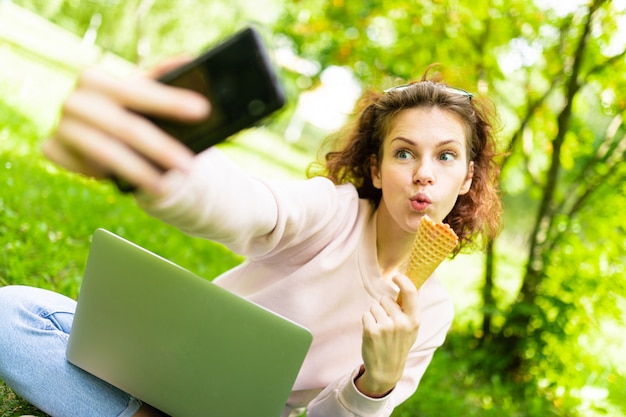 Jolie jeune femme de race blanche est engagée à la pige dans le parc avec un ordinateur portable, la tasse de café et de crème glacée, est assise sur l'herbe et fait selfie