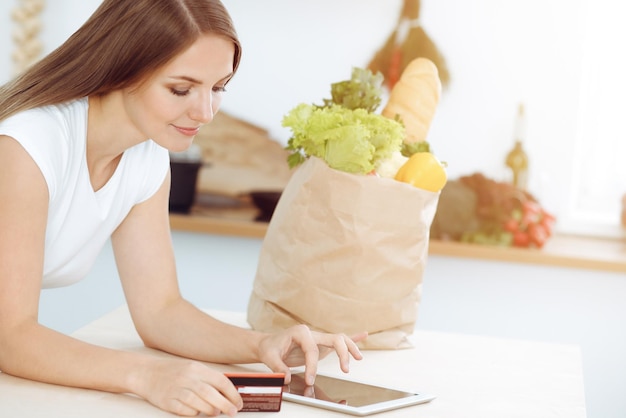 Une jolie jeune femme à la recherche d'une nouvelle recette pour un délicieux mélange de salade tout en étant assis à la table dans une cuisine ensoleillée Tablet pc est le meilleur livre de cuisine