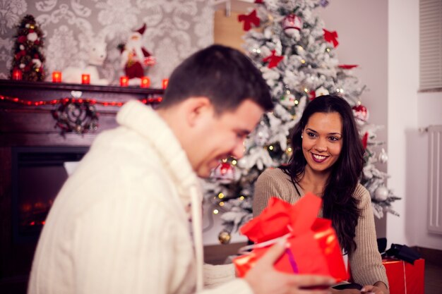 Jolie jeune femme regardant avec enthousiasme son mari pendant qu'il ouvre le cadeau. Le jour de Noël. Couple heureux.