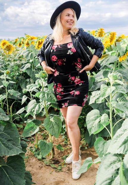 Jolie jeune femme en robe veste en cuir noir et chapeau pose dans un champ de tournesols