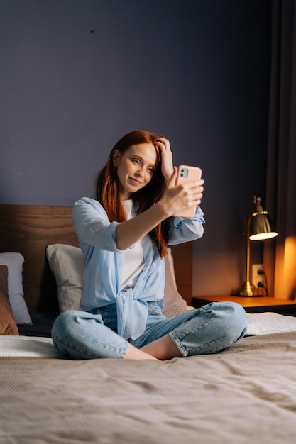 Jolie jeune femme rousse posant pour un selfie mobile au lit, assise sur le lit dans une chambre confortable. Femme de race blanche ayant des appels vidéo dans l'application de téléphonie mobile à la maison.