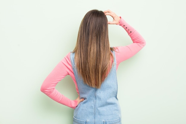 Jolie jeune femme se sentant désemparée et confuse en pensant à une solution avec la main sur la hanche et l'autre sur la vue arrière de la tête