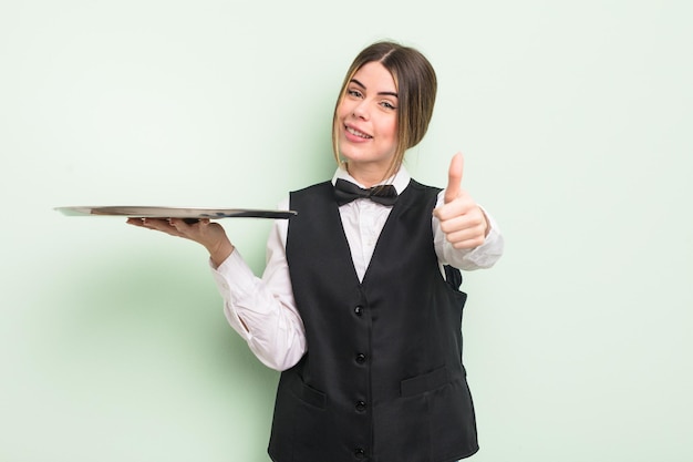 Jolie jeune femme se sentant fière, souriant positivement avec les pouces vers le haut. serveur avec un concept de plateau