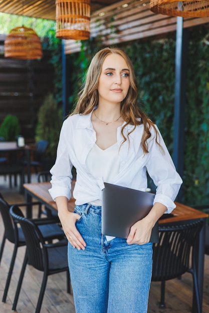 Une jolie jeune femme se tient sur une terrasse ouverte dans un café avec un ordinateur portable dans les mains Une femme en vêtements décontractés travaille à distance sur un ordinateur portable