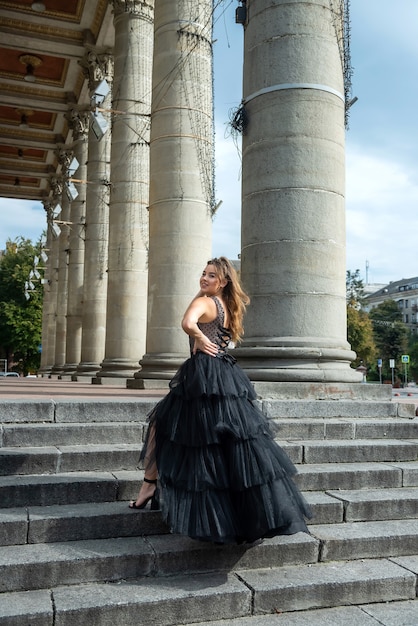 Jolie jeune femme sensuelle en robe de soirée cocktail noire posant près de l'entrée du théâtre de la ville avec des colonnes blanches