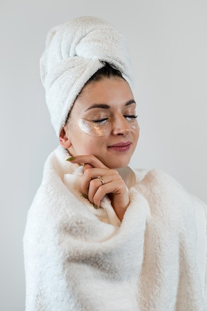 Jolie jeune femme en serviette blanche appliquant des patchs d'hydrogel de collagène sous les yeux pour lisser la peau isolée sur blanc