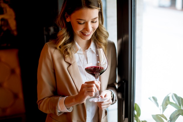 Jolie jeune femme seule avec un verre de vin rouge debout près de la fenêtre et regardant de côté