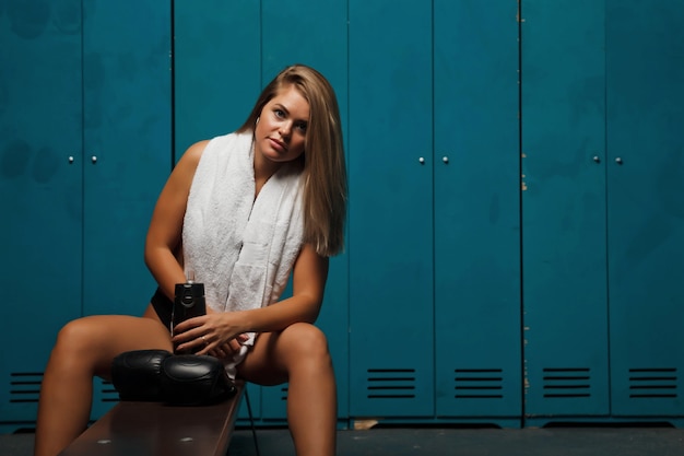 Jolie jeune femme avec une silhouette athlétique avec une bouteille d'eau dans un club de sport de vestiaire, avec une serviette et des gants de boxe