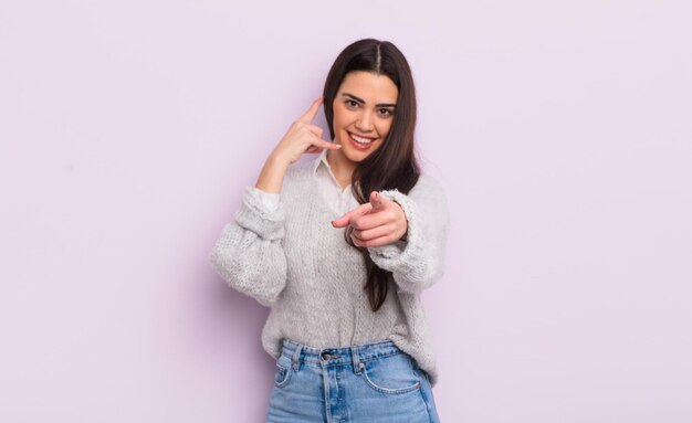 Jolie jeune femme souriant joyeusement et pointant vers la caméra tout en vous appelant plus tard, en parlant au téléphone