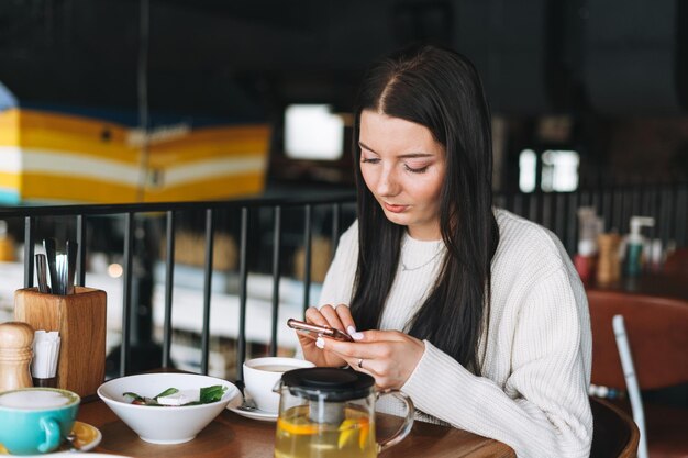 Jolie jeune femme souriante brune en décontracté à l'aide d'un téléphone portable ayant un brunch au café