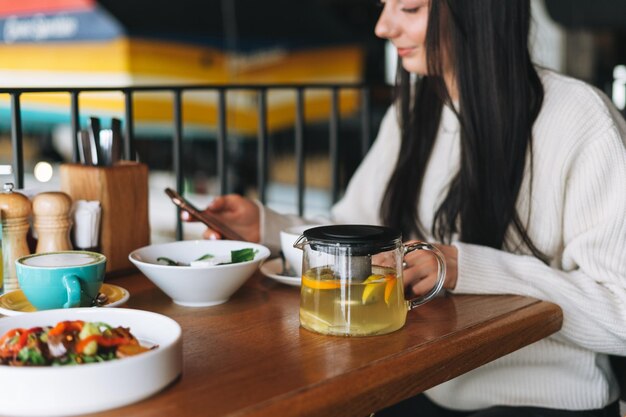 Jolie jeune femme souriante brune en décontracté à l'aide d'un téléphone portable ayant un brunch au café