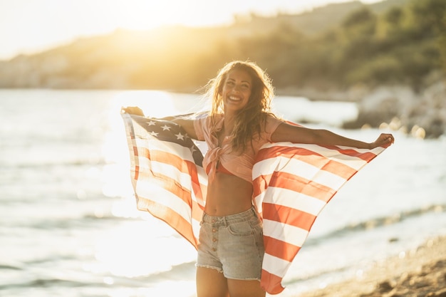 Jolie jeune femme souriante avec drapeau national américain profitant d'une journée de détente sur la plage.