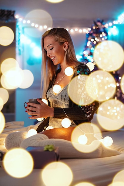 Jolie jeune femme souriante est assise sur le lit tenant une tasse de café, entourée de lumières à bulles de Noël.