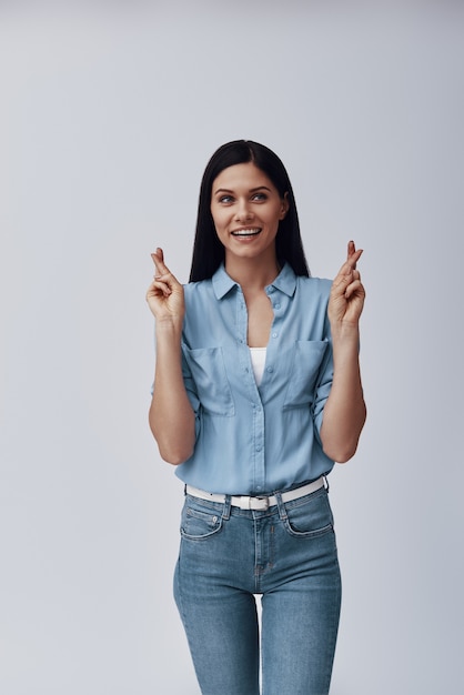 Jolie jeune femme souriante et gardant les doigts croisés en se tenant debout contre le mur gris