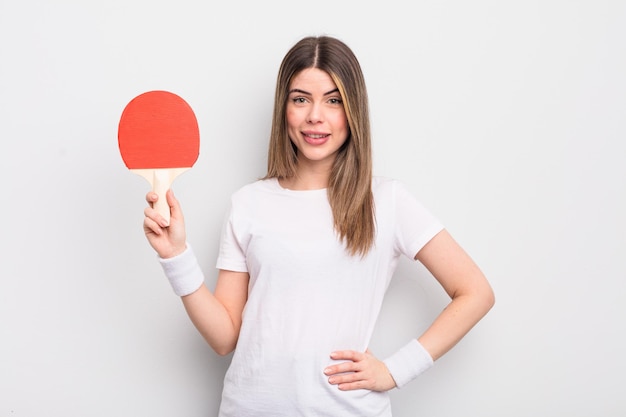 Jolie jeune femme souriante joyeusement avec une main sur la hanche et un concept de ping-pong confiant