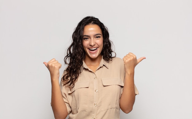 Jolie jeune femme souriante joyeusement et à la recherche de bonheur, se sentant insouciante et positive avec les deux pouces vers le haut