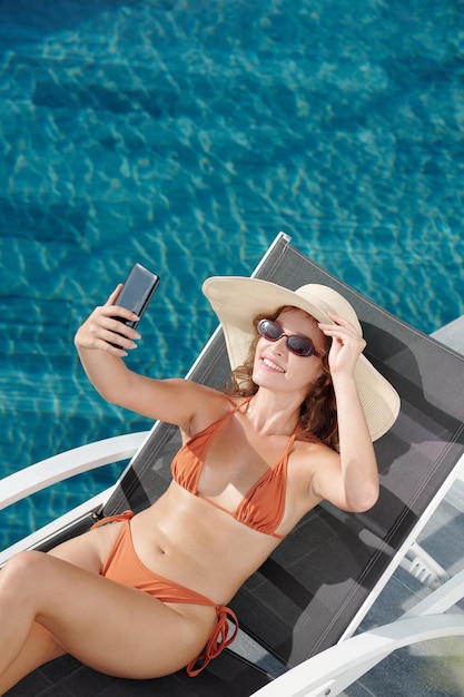 Jolie jeune femme souriante à lunettes de soleil et chapeau de paille prenant selfie au bord de la piscine