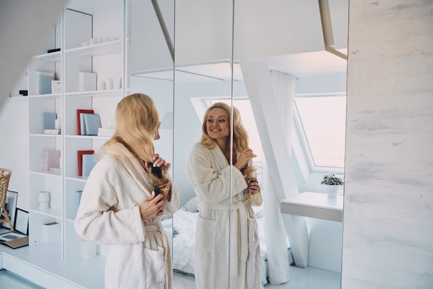 Jolie jeune femme souriante en peignoir peignant les cheveux tout en se tenant devant le miroir à la maison