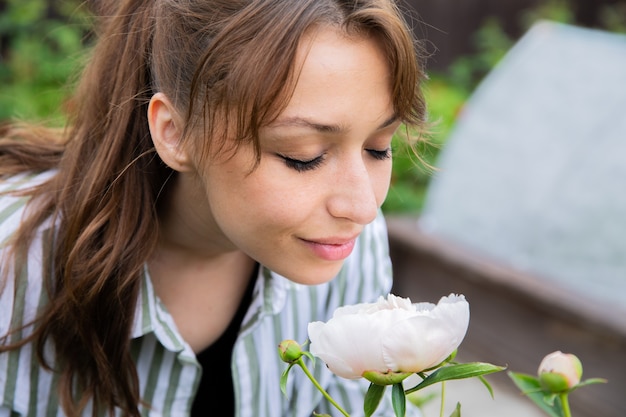 Jolie jeune femme souriante reniflant la pivoine rose dans le jardin de la maison de campagne