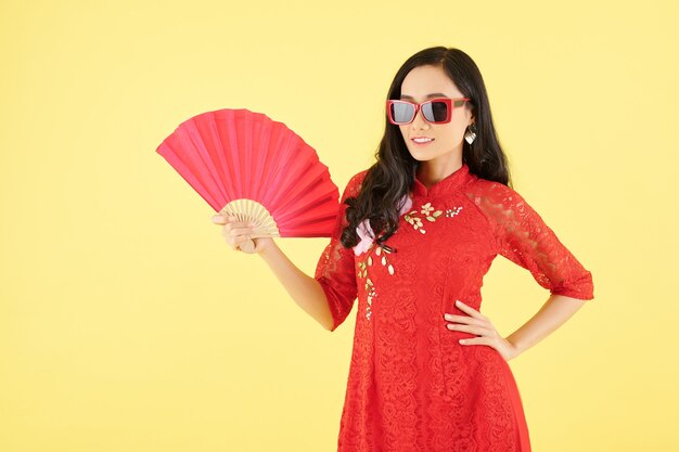 Jolie jeune femme souriante en robe rouge et lunettes de soleil standingwith fan de papier