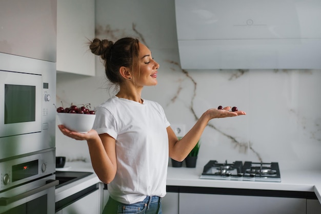 Jolie jeune femme souriante tient une assiette de baies de cerises fraîches dans sa main dans la cuisine