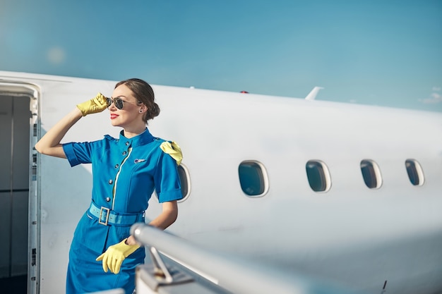 Une jolie jeune femme souriante en uniforme et lunettes de soleil se tient à la sortie de l'avion en attendant le départ