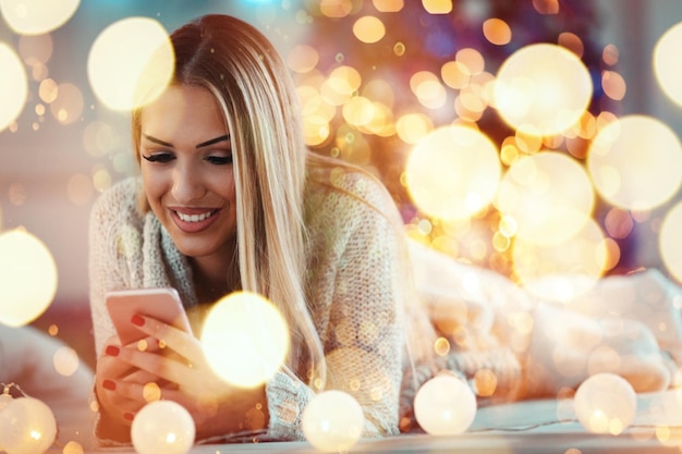 Jolie jeune femme souriante utilisant un smartphone et un sourire heureux pendant les vacances de Noël confortables à la maison.