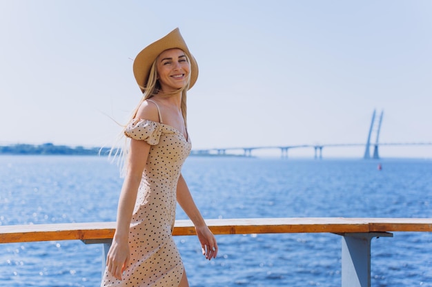 Jolie jeune femme suédoise blonde au chapeau marchant sur le remblai sur la baie de la mer et le grand pont en arrière-plan pendant les vacances d'été Maquette pour les voyages et les loisirs Mode de vie de luxe Belle fille