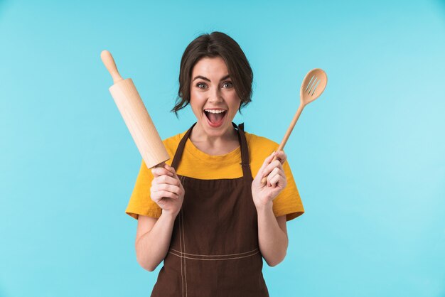 jolie jeune femme surprise excitée chef tenant un rouleau à pâtisserie et une cuillère en bois isolée sur un mur bleu.