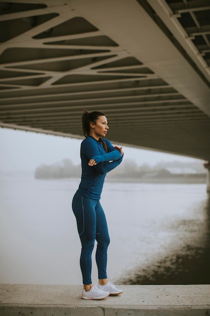 Jolie jeune femme en survêtement bleu s'étirant avant l'entraînement au bord de la rivière au matin d'automne