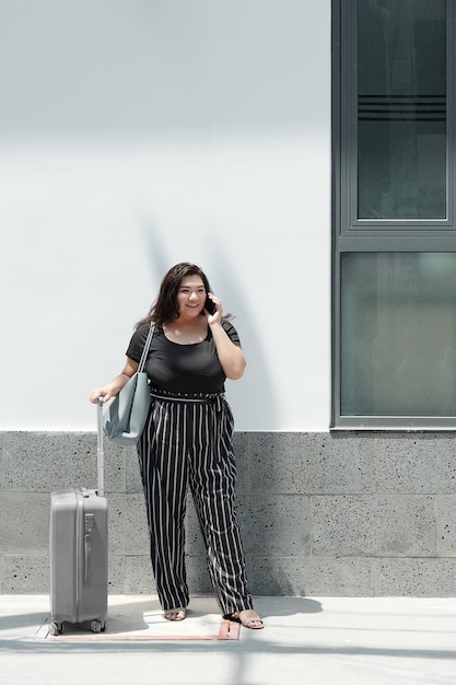 Jolie jeune femme de taille plus debout à l'extérieur avec une valise et un sac et parlant au téléphone avec un chauffeur de taxi