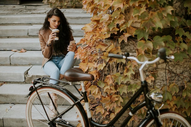 Jolie jeune femme avec téléphone portable boire du café à emporter dans les escaliers par le vélo le jour de l'automne