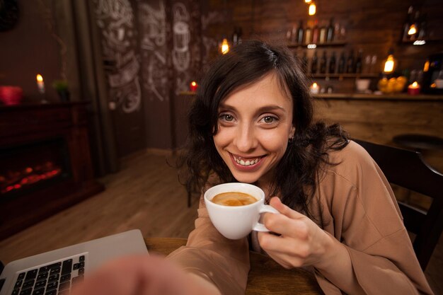 Jolie jeune femme tenant un appareil photo et une tasse de café prenant un selfie. Fille s'amusant dans un café. Jeune femme tenant une tasse de café.