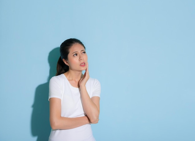 jolie jeune femme tenant sur le menton la performance de l'émotion inconfortable douloureuse de la dent et regardant la zone vide debout dans le fond du mur bleu.