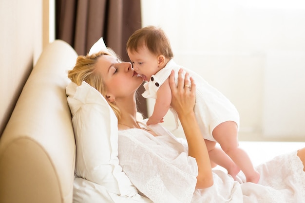 Jolie jeune femme tenant une petite fille dans ses bras à la chambre domestique le matin. Heureuse mère embrassant et embrassant sa petite fille nouveau-née à la maison sur un lit blanc