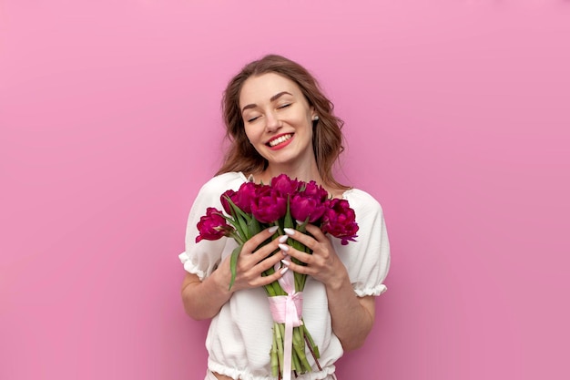 Une jolie jeune femme en tenue de fête tient un bouquet de tulipes roses et se réjouit sur un fond rose