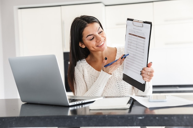 Jolie jeune femme travaillant avec un ordinateur portable et des documents alors qu'elle était assise à la cuisine, montrant un graphique