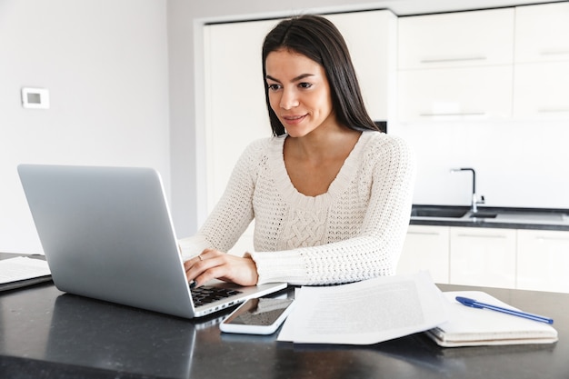 Jolie jeune femme travaillant avec un ordinateur portable et des documents alors qu'elle était assise à la cuisine