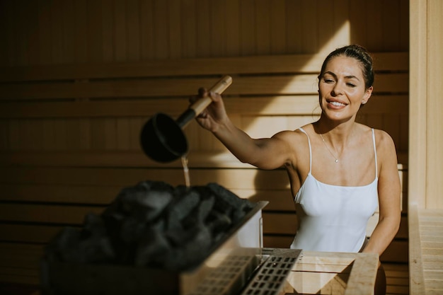 Jolie jeune femme versant de l'eau sur des pierres chaudes dans le sauna