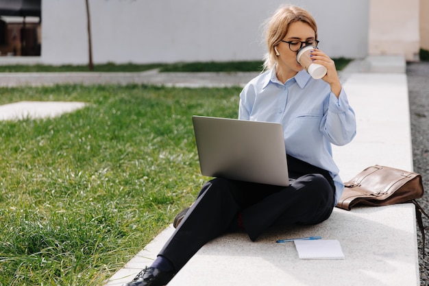 Jolie jeune femme en vêtements décontractés buvant du café frais et utilisant un ordinateur portable sans fil pour travailler à l'air frais. Concept de technologie indépendante et moderne.