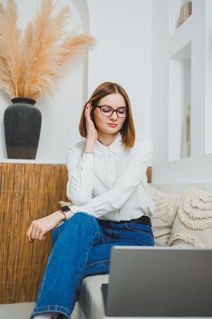 Une jolie jeune femme en vêtements décontractés est assise sur le canapé à la maison et travaille sur un ordinateur portable Travail en ligne à distance