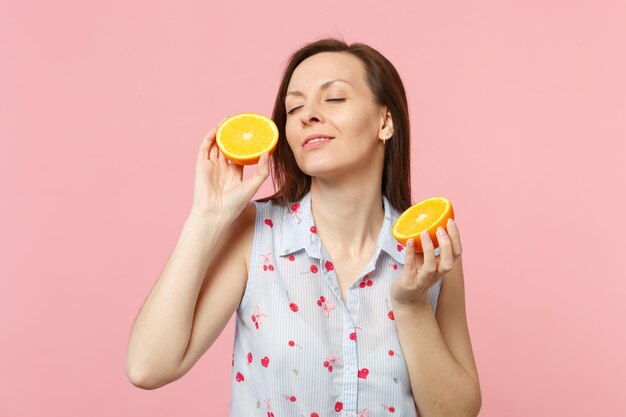 Jolie jeune femme en vêtements d'été gardant les yeux fermés tenant des moitiés de fruits orange mûrs frais isolés sur fond pastel rose. Les gens vivent un style de vie vivant et se détendent. Maquette de l'espace de copie.